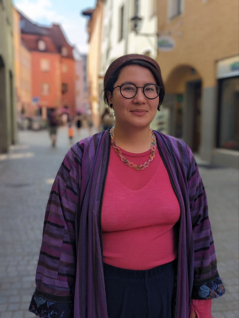 A woman with short dark hair wearing a Central Asian scullcap and a lila chapan coat standing in a cobblestone street with medieval buildings
