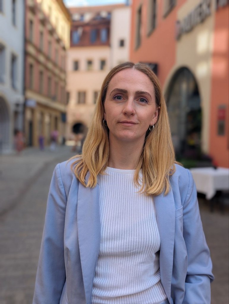 A blond young woman in a white top and a blue blazer standing in a cobblestone street with medieval buildings